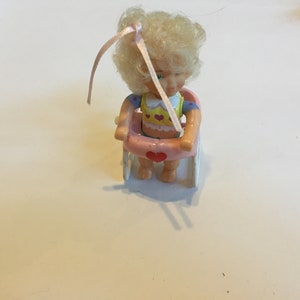 May include: A wind-up toy doll with blonde hair and a pink bow sits in a pink wheelchair with white wheels. The doll is wearing a yellow and blue outfit with hearts.