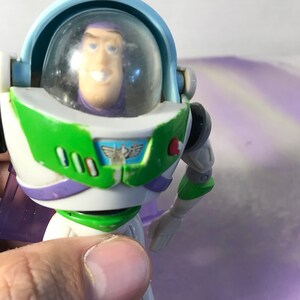 May include: A plastic toy action figure of Buzz Lightyear from the Toy Story franchise. The figure is wearing a white, green, and purple spacesuit with a clear plastic helmet. The figure has a red button on its chest and a green and white design on its chest.
