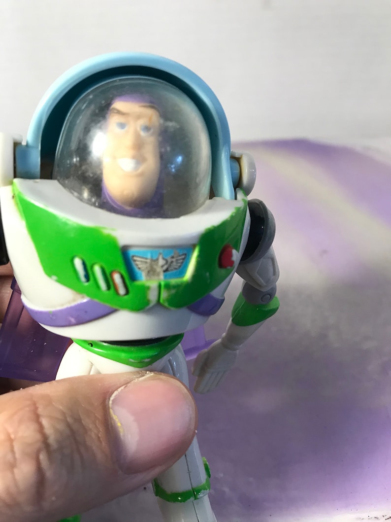 May include: A plastic toy action figure of Buzz Lightyear from the Toy Story franchise. The figure is wearing a white, green, and purple spacesuit with a clear plastic helmet. The figure has a red button on its chest and a green and white design on its chest.