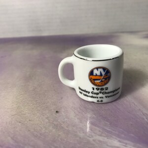 Może przedstawiać: Biały kubek ceramiczny z srebrną obwódką. Kubek przedstawia logo New York Islanders i tekst "1982 Stanley Cup® Champions NY Islanders vs. Vancouver 4-0".