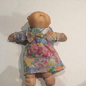 May include: A Cabbage Patch Kid doll wearing a floral dress with a blue bow. The doll has a light brown complexion and is lying on its back with its arms outstretched.