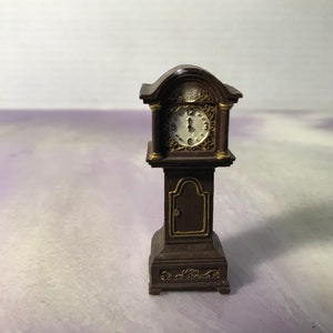 May include: A miniature brown grandfather clock with gold accents and a white clock face. The clock has a decorative design on the base and a door on the front.