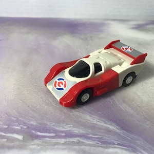 May include: A red, white, and gray toy race car with a black windshield. The car features a spoiler and a logo on the front and rear. The toy car is a vintage collectible.