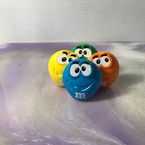 Könnte beinhalten: Vier bunte M&M's-Figuren mit lächelnden Gesichtern. Die Figuren sind gelb, grün, blau und orange.