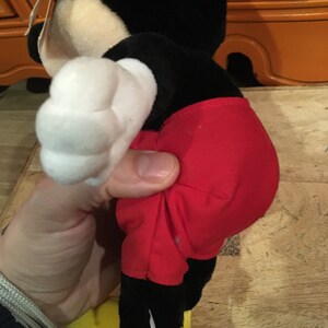 Może przedstawiać: Pluszowa zabawka Mickey Mouse w kolorze czarnym i czerwonym z białą rękawiczką. Zabawka stoi na drewnianej powierzchni.