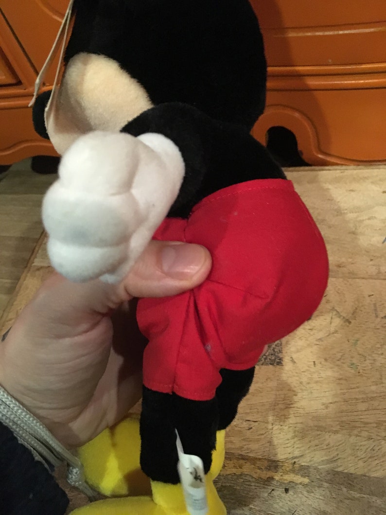 Może przedstawiać: Pluszowa zabawka Mickey Mouse w kolorze czarnym i czerwonym z białą rękawiczką. Zabawka stoi na drewnianej powierzchni.