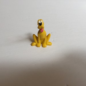 Pode incluir: Uma figurinha de plástico amarela de um cão de desenho animado com olhos pretos, uma língua rosa que se projeta e marcas brancas no peito. O cão está sentado com as pernas enfiadas debaixo do corpo.