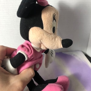 K&ouml;nnte beinhalten: Ein Pl&uuml;schtier von Minnie Mouse mit schwarzen Ohren, einer rosa Schleife und einem rosa Kleid mit wei&szlig;en Punkten. Das Spielzeug steht auf einer lila und wei&szlig;en Oberfl&auml;che.