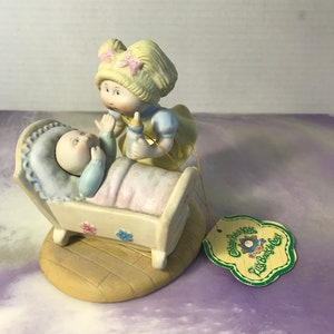 May include: A porcelain figurine of a Cabbage Patch Kid doll holding a baby doll in a cot. The doll is wearing a yellow dress with pink bows and the baby is wearing a blue outfit. The figurine has a tag that reads "Cabbage Patch Kids Little Buds of Love".
