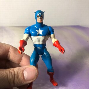 May include: A vintage Captain America action figure. The figure is primarily blue, with a white star on the chest and red gloves and boots. The figure has a blue mask and a stern expression.