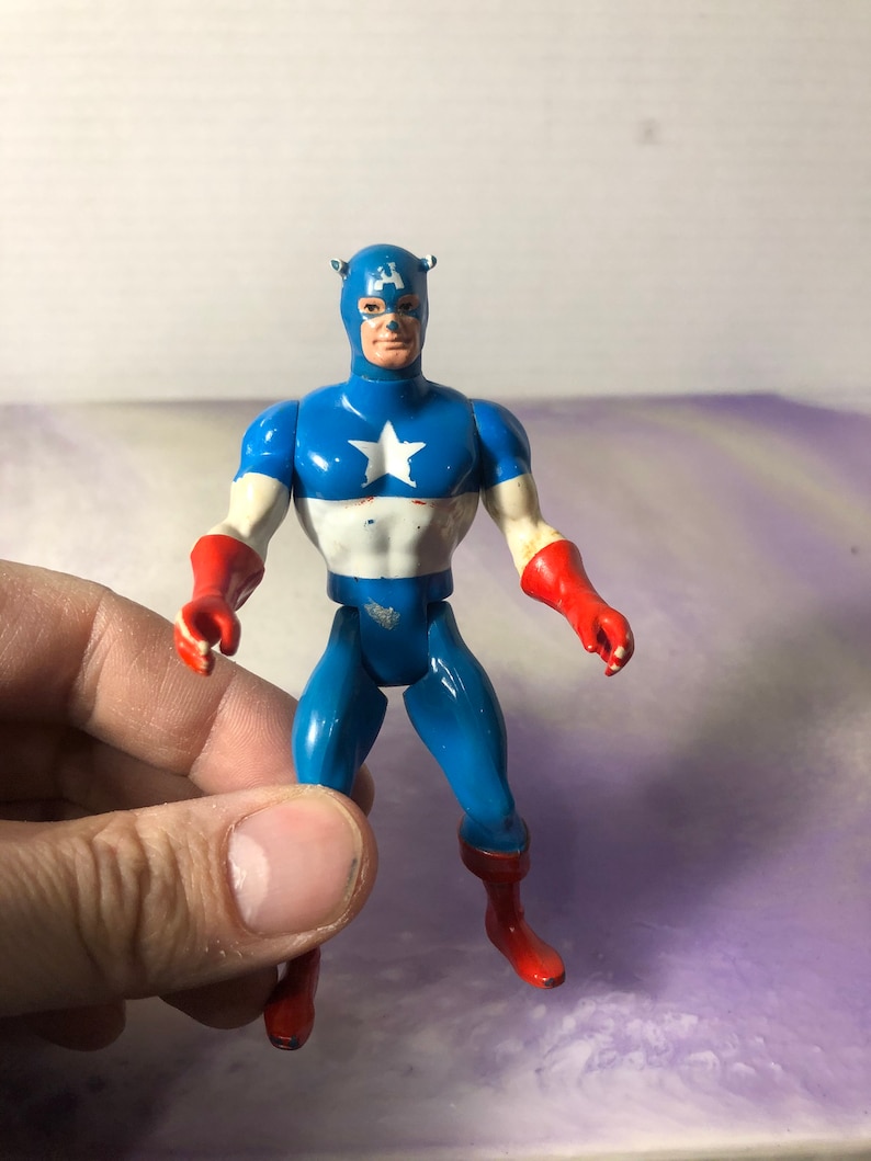 May include: A vintage Captain America action figure. The figure is primarily blue, with a white star on the chest and red gloves and boots. The figure has a blue mask and a stern expression.