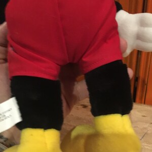 Puede incluir: Un juguete de peluche negro y rojo con zapatos amarillos. El juguete es un personaje de dibujos animados con una camisa roja y pantalones cortos negros.