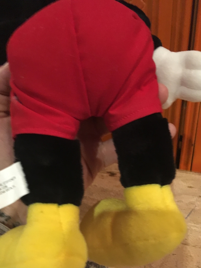 Puede incluir: Un juguete de peluche negro y rojo con zapatos amarillos. El juguete es un personaje de dibujos animados con una camisa roja y pantalones cortos negros.