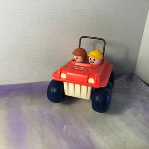 以下が含まれることがあります： 青い車輪と車内に2体のフィギュアが付いた赤いプラスチック製の玩具の車。車の前方に「Roaming Buggy」という文字が印刷されています。