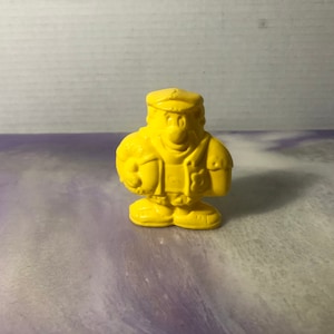 May include: A yellow plastic figurine of a cartoon character wearing a hat and a jacket. The character has a large smile and is holding a small object in its hand.