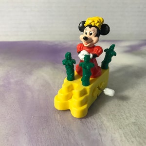 May include: A yellow wind-up toy featuring a red-dressed Mickey Mouse figure with a yellow crown. The toy has a yellow base with green palm trees.
