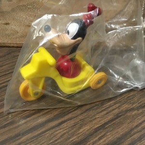May include: A yellow plastic toy tricycle with a cartoon character riding it. The character is black with a red hat and is smiling. The toy is in a clear plastic bag.