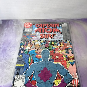 May include: A comic book cover featuring Captain Atom, a superhero with a blue and purple costume, standing in front of a group of other superheroes, including Superman, Batman, Wonder Woman, and The Flash. The cover text reads "Captain Atom Sir!" and "Injustice: First Strike!"