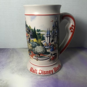 K&ouml;nnte beinhalten: Eine wei&szlig;e Keramiktasse mit einer bunten Illustration von Walt Disney World, einschlie&szlig;lich Cinderella's Castle, dem Magic Kingdom und anderen Attraktionen. Die Tasse hat einen roten Griff und die Worte "Walt Disney World" sind auf dem Boden aufgedruckt.