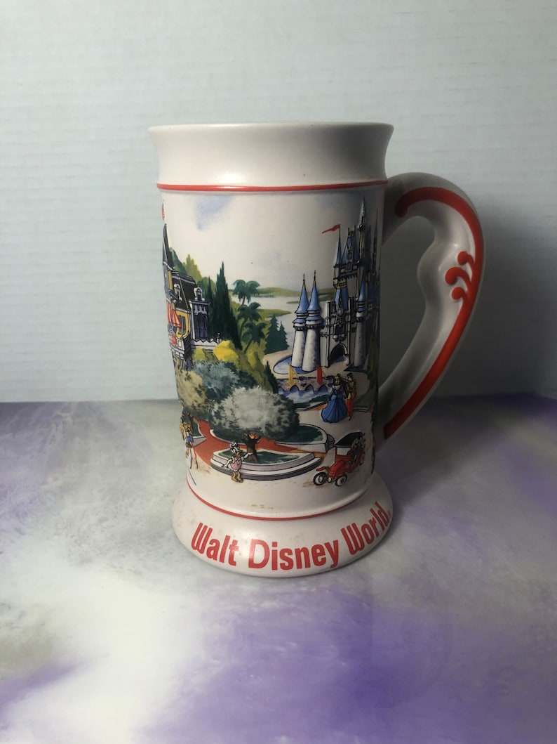 K&ouml;nnte beinhalten: Eine wei&szlig;e Keramiktasse mit einer bunten Illustration von Walt Disney World, einschlie&szlig;lich Cinderella's Castle, dem Magic Kingdom und anderen Attraktionen. Die Tasse hat einen roten Griff und die Worte "Walt Disney World" sind auf dem Boden aufgedruckt.