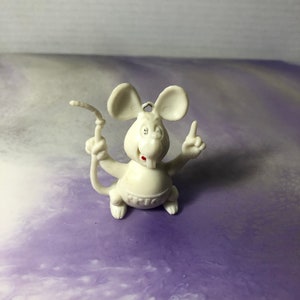 May include: White plastic figurine of a mouse with red eyes and a red mouth. The mouse is standing on its hind legs and holding its front paws up in the air. The mouse is wearing a white diaper with the word "MIC" written on it.