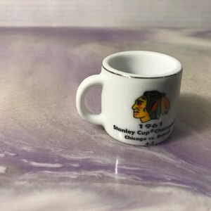 May include: White ceramic mug with a black and red Chicago Blackhawks logo and the text "1961 Stanley Cup Champions Chicago vs. Detroit 4-2".