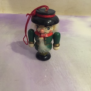 May include: A wooden nutcracker ornament with a black hat, green jacket, and white beard. The ornament has a red string for hanging.