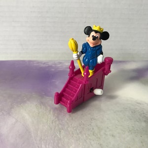 May include: A wind-up toy featuring Mickey Mouse dressed as a prince, wearing a blue and white outfit and a gold crown. He is holding a yellow scepter and standing on a pink castle-shaped platform with stairs.