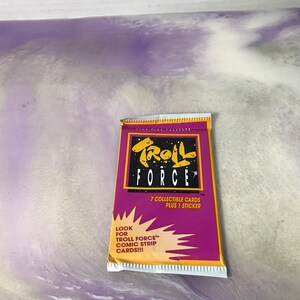 May include: A purple and yellow card pack for Troll Force trading cards. The pack contains 7 collectible cards and 1 sticker. The text on the pack reads "Troll Force" and "7 collectible cards plus 1 sticker".