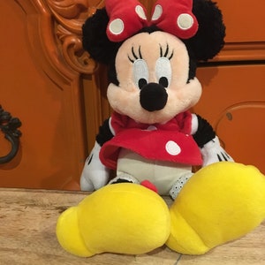 Op de afbeelding: Een pluche speelgoed van Minnie Mouse die een rode jurk draagt met witte stippen en een rode strik met witte stippen. Ze heeft zwarte oren en een zwarte neus. Ze zit op een houten oppervlak.