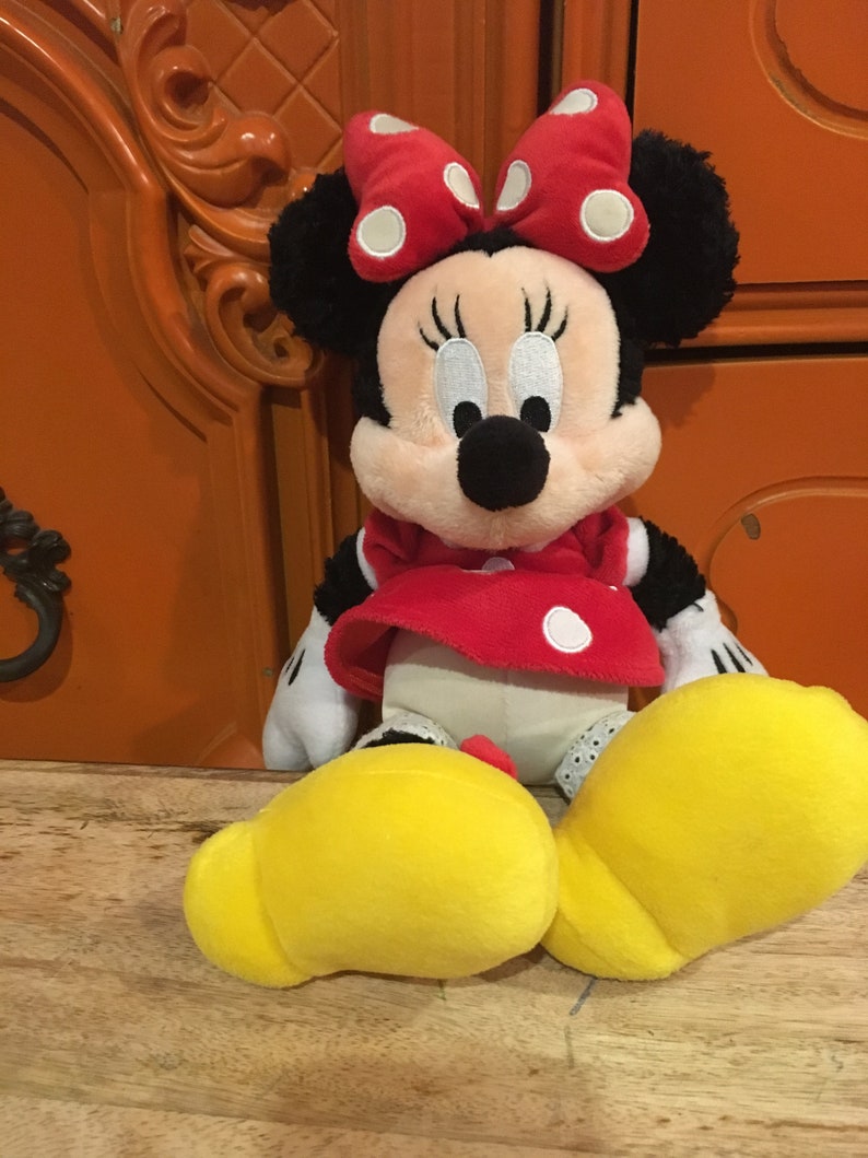 Op de afbeelding: Een pluche speelgoed van Minnie Mouse die een rode jurk draagt met witte stippen en een rode strik met witte stippen. Ze heeft zwarte oren en een zwarte neus. Ze zit op een houten oppervlak.