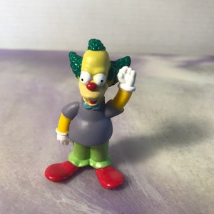 May include: A yellow Krusty the Clown figurine with green hair, a purple shirt, and red shoes. He is waving with his right hand.