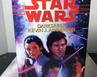 May include: A Star Wars paperback book cover featuring a purple, blue, and black space background. The title "STAR WARS" is in red and white. The book title "DARKSABER" is in white with the author's name "KEVIN J. ANDERSON" below it. The cover features a portrait of Luke Skywalker, Princess Leia, and Han Solo.