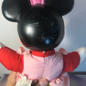 Op de afbeelding: Een pluche Minnie Mouse pop die een roze jurk met witte stippen, een rood jasje en een gele broek draagt. De pop heeft zwarte oren en een roze strik.