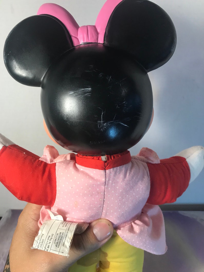 Op de afbeelding: Een pluche Minnie Mouse pop die een roze jurk met witte stippen, een rood jasje en een gele broek draagt. De pop heeft zwarte oren en een roze strik.