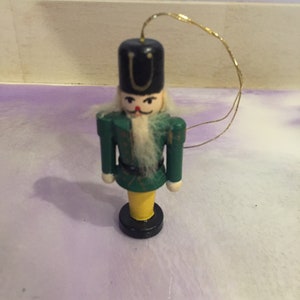 May include: A small wooden nutcracker ornament with a green and yellow painted design. The nutcracker is wearing a black hat with a gold trim and a white beard.