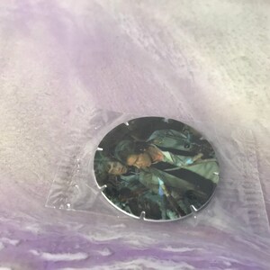 Può includere: Una moneta rotonda di colore argentato con un'immagine in bianco e nero di due persone. L'immagine è di un uomo e una donna, entrambi con i capelli scuri. L'uomo indossa una giacca marrone e la donna una camicia bianca. La moneta ha un bordo seghettato.