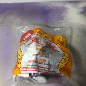 May include: A plastic bag containing a grey and white plush toy gopher. The bag is yellow and orange with the text "GOPHER SOFT TOY TAUPE JOUET SOUPLE GOFER MUÑEQUITO SUAVE" printed on it. See back for safety information. Instructions touchant la sécurité au dos du sac. Véase la información sobre seguridad al dorso.