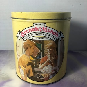 May include: A yellow metal tin with a picture of a boy and girl on the front. The tin is labeled "Franklin Crunch 'n Munch Buttery Toffee Popcorn with Peanuts".