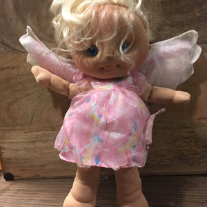 May include: A plush angel doll with blonde hair, blue eyes, and a pink dress with a sparkly design. The doll has white wings and is standing on a wooden surface.