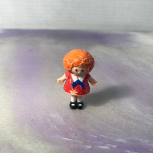 May include: A small plastic doll with red hair, wearing a red dress with a blue collar and bow. The doll is standing on a purple and white surface.