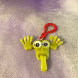 May include: Yellow plastic keychain with a cartoon face, googly eyes, and outstretched arms. The keychain has a red plastic clip.