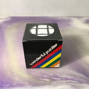 May include: A black box with the words "wonderful puzzler" in white, with a rainbow stripe design. The top of the box has a circular window showing a white puzzle. A classic brain teaser.