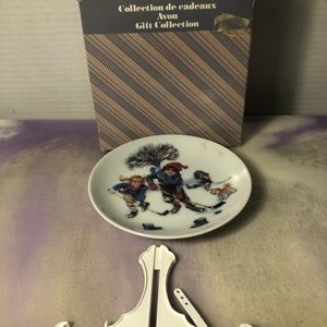 May include: A white ceramic plate featuring a winter scene of children ice skating. The plate is displayed on a white plastic stand. The plate is part of the Avon Winter Wonderland Mini Plate Series, Breaking the Ice. The box for the plate is visible in the background.