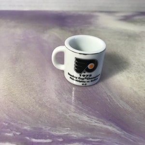 May include: White ceramic mug featuring a black and orange Philadelphia Flyers logo. The mug reads "1975 Stanley Cup Champions Philadelphia vs. Buffalo 4-2".