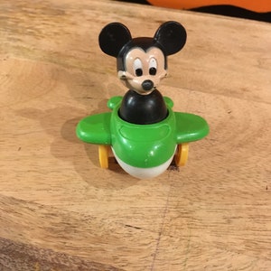 May include: A green toy aeroplane with a Mickey Mouse figure in the cockpit. The aeroplane has yellow wheels and a white propeller.