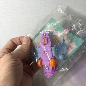 May include: Purple and blue plastic toy bat with orange wheels. The toy is in a clear plastic bag with a green and white McDonald's Happy Meal wrapper.