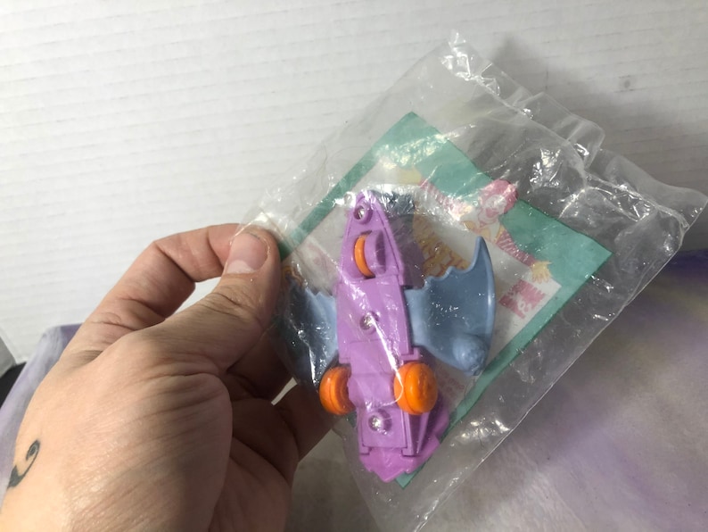 May include: Purple and blue plastic toy bat with orange wheels. The toy is in a clear plastic bag with a green and white McDonald's Happy Meal wrapper.