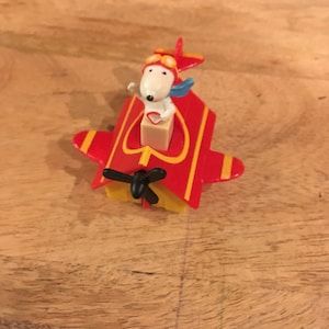 Op de afbeelding: Een rood en geel speelgoedvliegtuig met een witte hond die een bril en een blauwe sjaal in de cockpit draagt. Het vliegtuig heeft een propeller en een hartvormig raam.