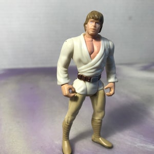 May include: A white plastic action figure of Luke Skywalker from Star Wars. He is wearing a white robe and brown pants. He has brown hair and blue eyes.